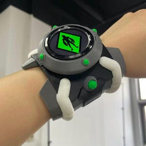 Può includere: Un orologio Omnitrix grigio e nero con dettagli verdi e uno schermo verde che mostra una silhouette. L'orologio è attaccato a un cinturino grigio e nero con dettagli bianchi.
