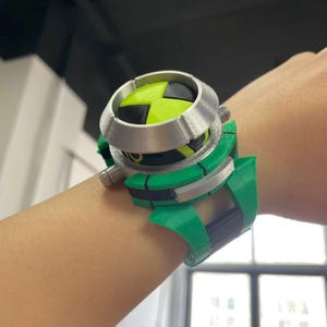 May include: A green and silver toy watch with a black and yellow face is worn on a wrist. The watch has a circular face with a four-way arrow design. The watch band is green with black accents.