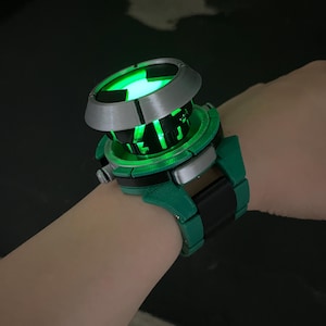 May include: A wrist-worn device featuring a circular, metallic silver top section with a glowing green light emanating from the centre. The base is constructed from green and black segments, forming a thick, mechanical-style band around the wrist.