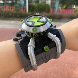 May include: A wrist-worn device featuring a black and silver frame with a circular top dial. The dial displays a bright green hourglass symbol. Three small, translucent green circular accents are embedded around the silver bezel and base.