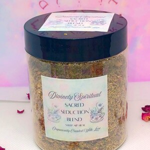 May include: A transparent jar with a black lid containing a herbal blend. The label says "Divinely Spiritual Sacred Seduction Blend." The background is pink with a floral design and scattered rose petals.