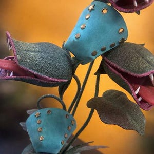 May include: A whimsical, handmade sculpture of a Venus flytrap-like plant. The plant features teal-colored, riveted pods and open, toothy mouths with red tongues. The leaves are dark green, and the background is a warm yellow.