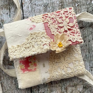 SMALL HAND STITCHED NECKLACE
Beautifully hand stitched pouch to hold your precious memories, a photograph or crystals. Made by slow stitching vintage French linens, lace, silk and quilting fabrics in shades of cream and white, lined with a pretty pink vintage paisley cotton, embellished with small silver bugle beads and a vintage fabric flower. 
It has a simple fold over top with a raw edged cream sari silk ribbon to wear around the neck.
Pouch: 3+1/4" / 8 cm square.
Ribbon drop: 16+1/2" / 42 cm