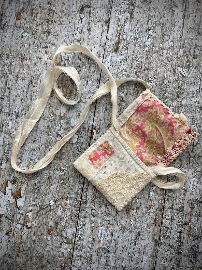 SMALL HAND STITCHED NECKLACE
Beautifully hand stitched pouch to hold your precious memories, a photograph or crystals. Made by slow stitching vintage French linens, lace, silk and quilting fabrics in shades of cream and white, lined with a pretty pink vintage paisley cotton, embellished with small silver bugle beads and a vintage fabric flower. 
It has a simple fold over top with a raw edged cream sari silk ribbon to wear around the neck.
Pouch: 3+1/4" / 8 cm square.
Ribbon drop: 16+1/2" / 42 cm