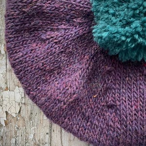 May include: A close-up of a hand-knitted hat in shades of purple with a teal pom-pom. The hat has a textured appearance, with visible stitches and a soft, fuzzy texture. The pom-pom is round and fluffy, adding a playful element.