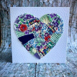 May include: A white card featuring a heart-shaped patchwork design crafted from colourful fabric scraps. The heart incorporates a variety of floral patterns in shades of blue, green, purple, and red.