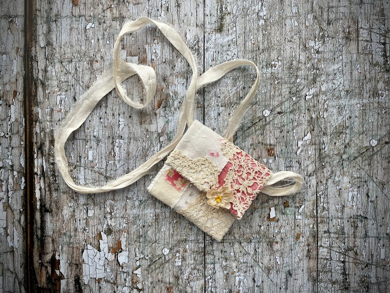SMALL HAND STITCHED NECKLACE
Beautifully hand stitched pouch to hold your precious memories, a photograph or crystals. Made by slow stitching vintage French linens, lace, silk and quilting fabrics in shades of cream and white, lined with a pretty pink vintage paisley cotton, embellished with small silver bugle beads and a vintage fabric flower. 
It has a simple fold over top with a raw edged cream sari silk ribbon to wear around the neck.
Pouch: 3+1/4" / 8 cm square.
Ribbon drop: 16+1/2" / 42 cm