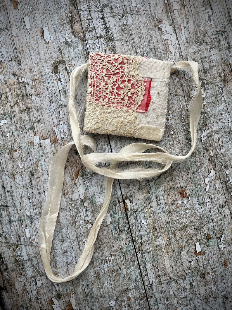 SMALL HAND STITCHED NECKLACE
Beautifully hand stitched pouch to hold your precious memories, a photograph or crystals. Made by slow stitching vintage French linens, lace, silk and quilting fabrics in shades of cream and white, lined with a pretty pink vintage paisley cotton, embellished with small silver bugle beads and a vintage fabric flower. 
It has a simple fold over top with a raw edged cream sari silk ribbon to wear around the neck.
Pouch: 3+1/4" / 8 cm square.
Ribbon drop: 16+1/2" / 42 cm