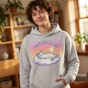 Può includere: Felpa con cappuccio grigia chiara con una grafica di un gatto che dorme su una nuvola e la scritta "Lost in the Moment". Il design include una farfalla e una palette di colori pastello.