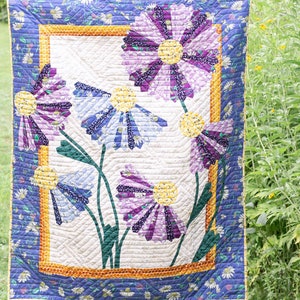 May include: A quilt featuring a floral design with purple, blue, and yellow flowers. The quilt is called "Dresdens in Bloom" and is designed by Rachel Rossi.