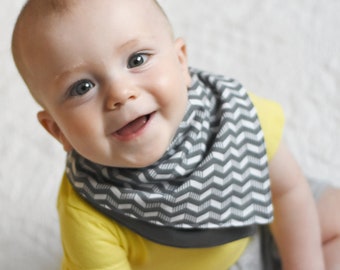 May include: A baby wearing a yellow shirt and a gray and white chevron bib. The baby is smiling and looking up at the camera.