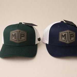May include: Two trucker hats with white mesh backs and solid front panels. The left hat is dark green, and the right hat is navy blue. Each features a centered hexagonal patch with the letters MP in a bold, dark font and a black tag attached.