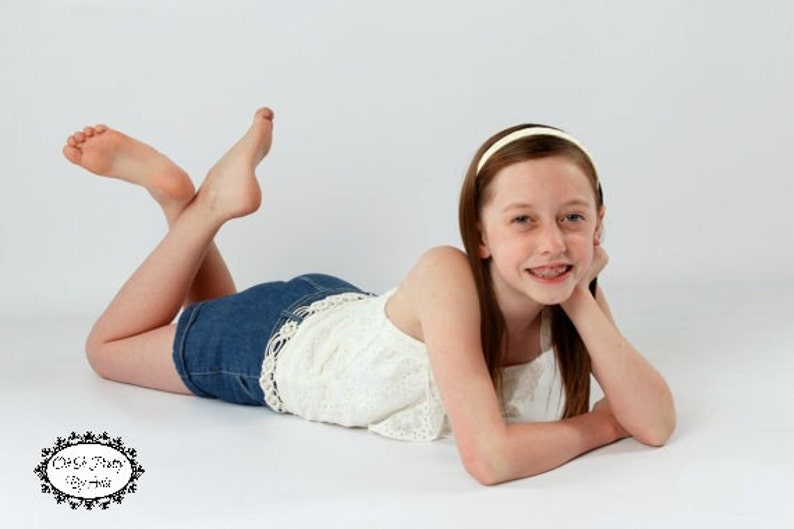 Pode incluir: Uma menina jovem com cabelo castanho comprido, vestindo um top branco sem mangas e shorts jeans azuis, est&aacute; deitada de bru&ccedil;os em um fundo branco. Ela est&aacute; sorrindo e tem aparelho nos dentes. Ela est&aacute; usando uma faixa de cabe&ccedil;a branca.