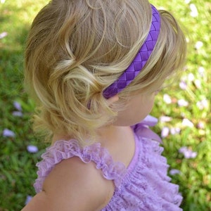 May include: A young girl with blonde hair wearing a purple braided headband and a lavender lace dress.