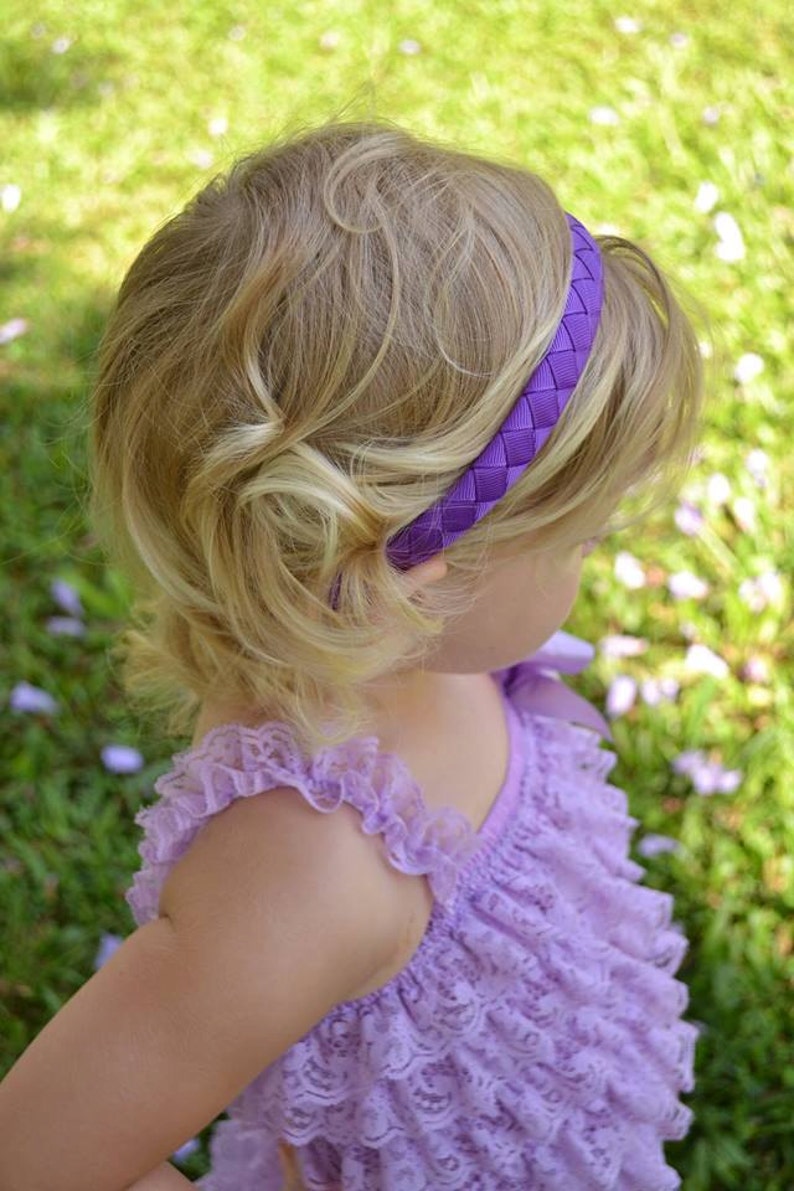 May include: A young girl with blonde hair wearing a purple braided headband and a lavender lace dress.