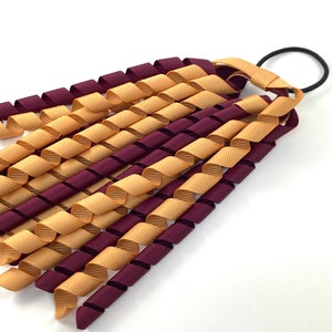 May include: A burgundy and gold hair scrunchie with a curled ribbon design. The scrunchie is made of two colours of ribbon that are twisted together.