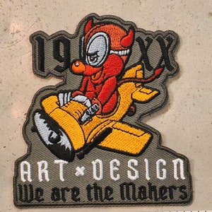 Könnte beinhalten: Ein grauer gestickter Aufnäher mit einer roten Teufelsfigur, die einen Helm trägt und ein gelbes Flugzeug fliegt. Der Text "ARTDESIGN We are the Makers" ist in Weiß auf den Aufnäher gestickt.