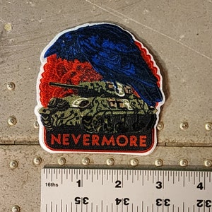 May include: A green military tank with a red, white, and blue crow perched on top. The tank is surrounded by red flames. The words "NEVERMORE" are printed in black on a white background below the tank.