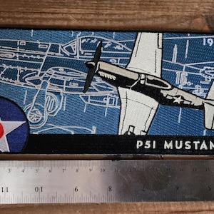 May include: Embroidered patch featuring a P51 Mustang aircraft design. The patch has a blue background with a white outline of the plane and a circular emblem with a white star and red center. The text "P51 MUSTANG" is displayed.