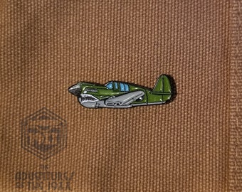 May include: Green enamel pin of a vintage airplane with a blue window. The pin is on a brown fabric background.