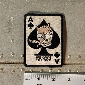 May include: A black and white embroidered patch of a playing card, the Ace of Spades. The card features a skull wearing a pilot's helmet and goggles. The text "WE OWN THE SKY" is printed below the card.