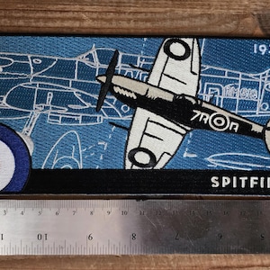 May include: Embroidered patch featuring a vintage Spitfire airplane design. The patch has a blue background with white outlines of the plane and a roundel. The word "SPITFIRE" is at the bottom. The patch measures approximately 8 inches long.