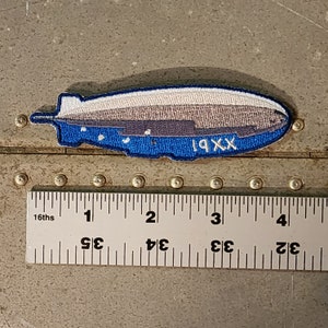 Könnte beinhalten: Ein blauer und grauer gestickter Aufnäher eines Luftschiffs mit dem Text "19XX" an der Seite. Der Aufnäher ist etwa 6,3 cm lang.