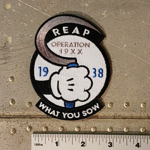 Peut inclure: Un écusson brodé noir et blanc avec une main de dessin animé tenant une clé. L'écusson porte le texte "REAP OPERATION 19XX 19 38 WHAT YOU SOW" en lettres blanches et brunes.