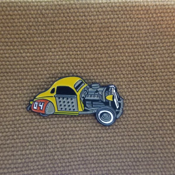 May include: A yellow and grey enamel pin of a vintage hot rod car with the number "04" on the side.