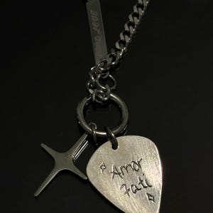 May include: A silver-toned necklace with a guitar pick-shaped pendant engraved with "Amor Fati" and a star-shaped charm. The necklace features a curb chain and a bar with the text "8:08 LOVE".