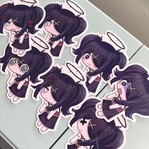 Puede incluir: Una colección de siete pegatinas con un personaje de dibujos animados con pelo oscuro, un halo y un gorro de enfermera. El personaje tiene varias expresiones faciales, incluyendo corazones, ojos cruzados y una cara enfadada. Las pegatinas están sobre una superficie verde claro.