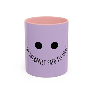 May include: A light purple ceramic mug with a soft pink interior rim. The front features two solid black circular eyes and the black text "MY THERAPIST SAID ITS OKAY" curved underneath, mimicking a simple, minimalist facial expression.