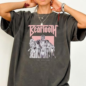 May include: Dark gray t-shirt featuring the band name "Beartooth" in pink block letters above a silver and pink graphic. The graphic depicts a figure with the text "The Surface" below.