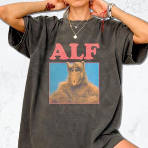 Puede incluir: Camiseta gris oscuro con la palabra "ALF" en rosa sobre una imagen rectangular del personaje ALF. La imagen de ALF está sobre un fondo azul.