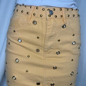 May include: A light orange denim mini skirt with gold-colored grommets and studs. The skirt has a classic five-pocket design and a button closure. The waistline and pockets are adorned with gold studs and grommets, adding a touch of edgy style.