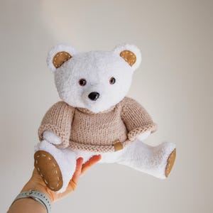 Könnte beinhalten: Ein weißer Teddybär mit einem beigefarbenen Pullover und braunen Akzenten an Ohren und Füßen. Der Bär hat braune Augen und eine schwarze Nase. Der Pullover hat einen lederartigen Aufnäher. Ein Plüschtier.