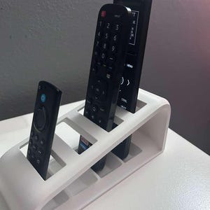 May include: A white plastic remote control organizer holding three black remote controls. The organizer has a curved design with multiple slots to hold the remotes upright. The remotes have various buttons and labels.