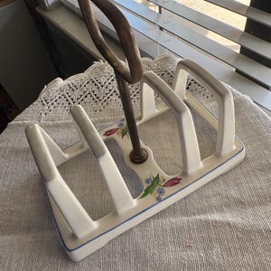 May include: Vintage white ceramic toast rack with a blue trim and floral accents. Features a metal handle and slots for multiple slices of toast. A charming kitchenware item.