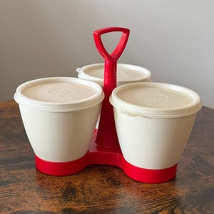 Puede incluir: Un especiero vintage rojo y crema. Cuenta con tres recipientes individuales de color crema con tapas blancas, colocados sobre una base roja. Un asa roja conecta los recipientes.