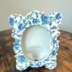 Puede incluir: Un marco de fotos de cerámica blanca con una abertura central ovalada y un borde festoneado. El marco está decorado con un patrón repetitivo de rosas azules, hojas verdes y pequeños detalles florales azules en toda la superficie de cerámica.