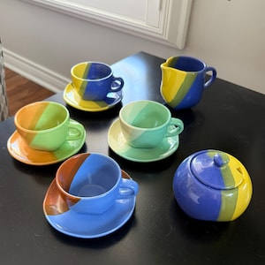 Peut inclure: Un ensemble de six tasses et soucoupes en céramique colorées, ainsi qu'une crémière et un sucrier. Les tasses et soucoupes présentent des rayures diagonales bleues, jaunes, vertes et oranges. Le sucrier a un couvercle. L'ensemble est disposé sur une surface noire.