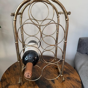 Peut inclure: Un casier à vin en métal doré avec une structure inspirée du bambou et six supports circulaires pour bouteilles. Une bouteille de vin en verre foncé avec une capsule cuivrée est placée dans l'emplacement circulaire inférieur gauche.