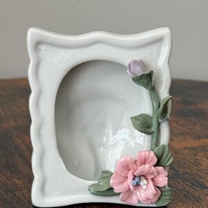 Könnte beinhalten: Ein weißer Keramik-Bilderrahmen mit floralem Design. Der Rahmen hat einen gewellten Rand und zeigt eine rosa Blume, eine lila Knospe und grüne Blätter. Der Rahmen steht auf einer Holzoberfläche.