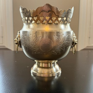 Puede incluir: Un cuenco de metal plateado con un patrón floral decorativo y asas con cabeza de león. El cuenco tiene una base de pedestal y un borde festoneado con recortes en forma de corazón. Probablemente vintage o antiguo.