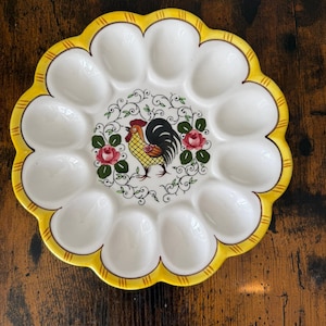 May include: A white ceramic egg plate with a yellow scalloped edge and a rooster design in the centre. The plate has 12 egg-shaped indentations and is decorated with floral accents. The rooster is yellow, black, and red.