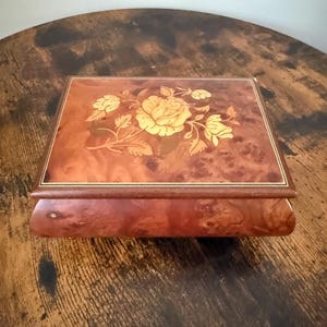 May include: A rectangular wooden music box featuring a polished burl wood finish with a light-coloured floral inlay on the lid. The top surface displays a detailed rose design crafted from wood veneer, bordered by thin, light-coloured decorative lines.