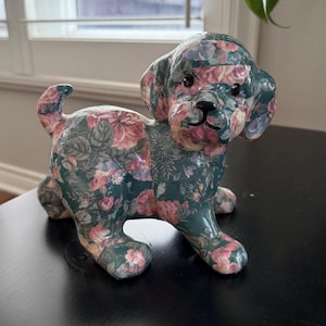 May include: A ceramic dog figurine with a floral pattern in pink, blue, and white on a dark green background. The dog is in a sitting position with its tail curled up. The figurine is on a black surface.