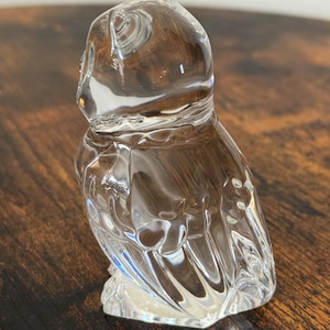 May include: A clear glass figurine shaped like an owl, featuring smooth, rounded contours and molded details that define the head, eyes, and folded wings. The transparent material reflects light, highlighting the sculpted form of the bird.