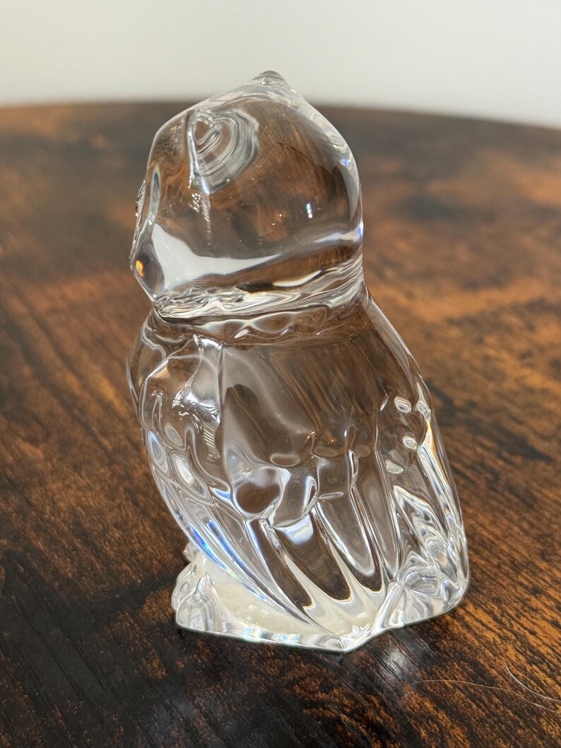 May include: A clear glass figurine shaped like an owl, featuring smooth, rounded contours and molded details that define the head, eyes, and folded wings. The transparent material reflects light, highlighting the sculpted form of the bird.
