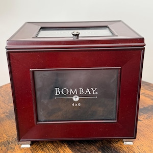 May include: A dark cherry-finished wooden box with a hinged lid featuring a glass display window. The front panel includes a rectangular frame with the text BOMBAY and 4 x 6 printed on the glass. The box has a small silver knob and silver feet.